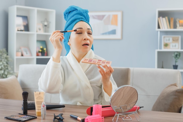 Jeune femme asiatique heureuse souriante avec une serviette sur la tête assise à la coiffeuse à l'intérieur de la maison en appliquant des fards à paupières en regardant le miroir faire la routine de maquillage du matin