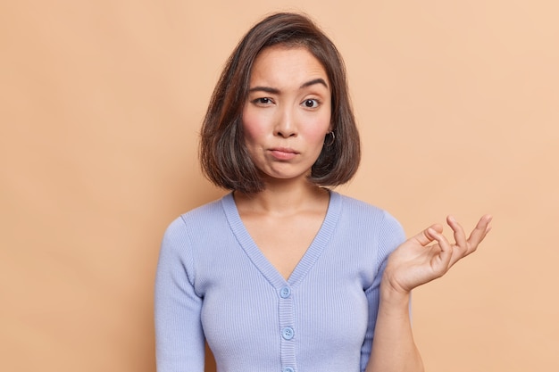 Une jeune femme asiatique ignorante lève la paume avec hésitation ne peut pas décider quoi faire semble incertaine vêtue d'un pull bleu décontracté isolé sur un mur beige a des doutes a une expression confuse