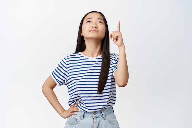 Jeune femme asiatique incertaine pointant vers le haut avec troublé sur blanc