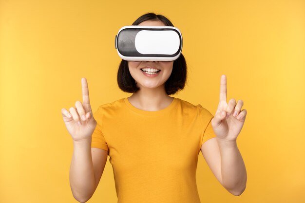 Jeune femme asiatique jouant en VR à l'aide de lunettes de réalité virtuelle portant un t-shirt jaune sur fond de studio