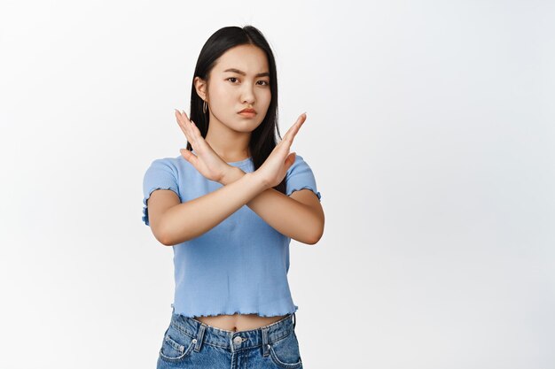 Jeune femme asiatique montre un geste de bloc d'arrêt croisé dire non interdire quelque chose de mauvais fronçant les sourcils mécontent debout sur fond blanc