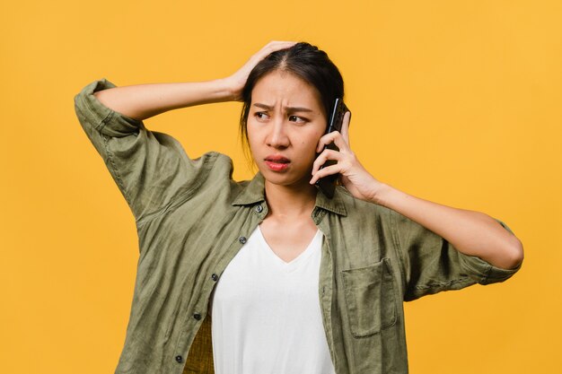 Une jeune femme asiatique parle par téléphone avec une expression négative, des cris excités, des cris émotionnels en colère dans un tissu décontracté et se tient isolée sur un mur jaune avec un espace de copie vierge. Concept d'expression faciale.