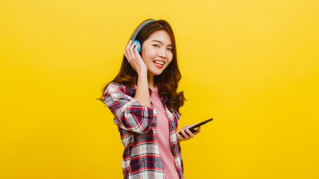 Jeune femme asiatique portant des écouteurs sans fil, écoutant de la musique de smartphone avec une expression joyeuse dans des vêtements décontractés et regardant la caméra sur le mur jaune. Concept d'expression faciale.