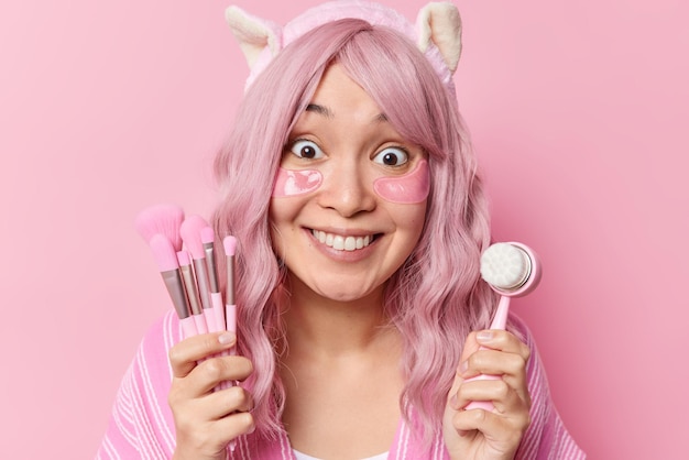 Une jeune femme asiatique positive avec une expression heureuse tient des pinceaux cosmétiques et un masseur de visage applique des patchs d'hydrogel a des cheveux teints isolés sur fond rose. Soins de la peau et traitements de bien-être