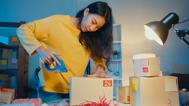 Jeune femme asiatique préparant le produit à l'aide de la boîte d'emballage de bande pour envoyer au client le bon de commande au bureau à domicile pendant la nuit