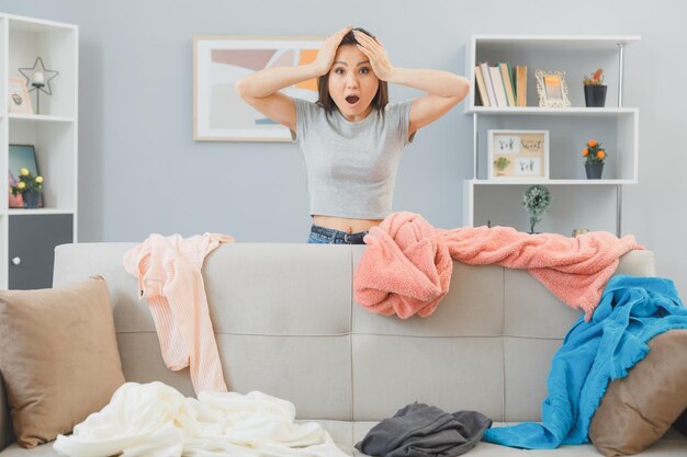 Jeune femme asiatique regardant le désordre dans son salon à la lessive dispersée dans la panique tenant les mains sur la tête étant inquiète et stressée