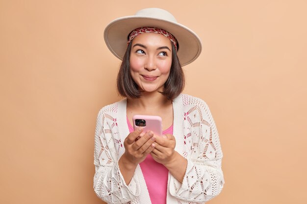 Une jeune femme asiatique rêveuse et ravie tient un smartphone moderne dans les mains utilise une application cellulaire pour discuter en ligne regarde ailleurs porte un chapeau élégant, un châle tricoté blanc isolé sur un mur marron