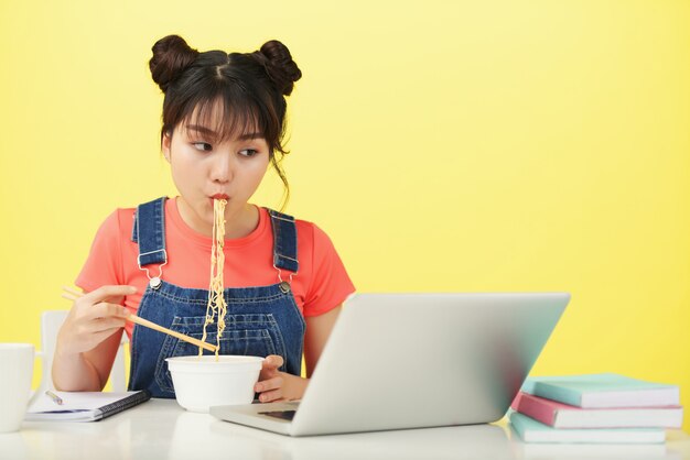 Jeune, femme asiatique, séance, bureau, manger, pot, nouilles, à, baguettes, et, regarder, ordinateur portable