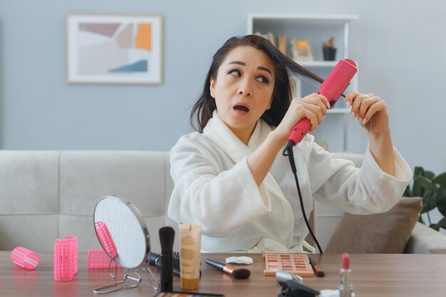 Jeune femme asiatique avec une serviette en peignoir assis à la coiffeuse à l'intérieur de la maison à l'aide d'un fer à friser rendant le style confus en faisant la routine de maquillage du matin