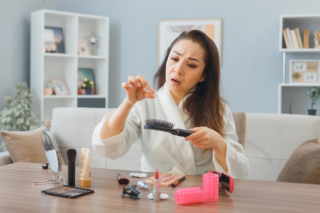 Jeune femme asiatique avec une serviette en peignoir assis à la coiffeuse à l'intérieur de la maison se brossant les cheveux étant confus et bouleversé d'avoir perdu les cheveux en faisant la routine de maquillage du matin