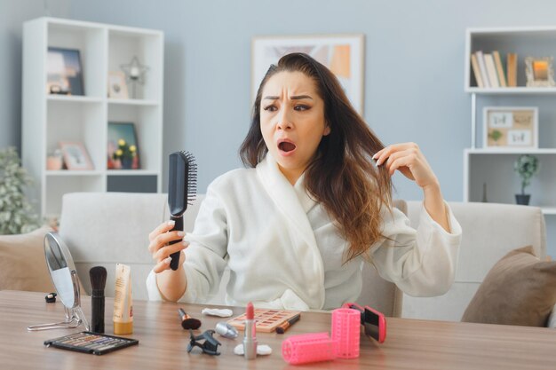 jeune femme asiatique avec une serviette en peignoir assis à la coiffeuse à l'intérieur de la maison se brosser les cheveux étant confus et contrarié de faire la routine de maquillage du matin