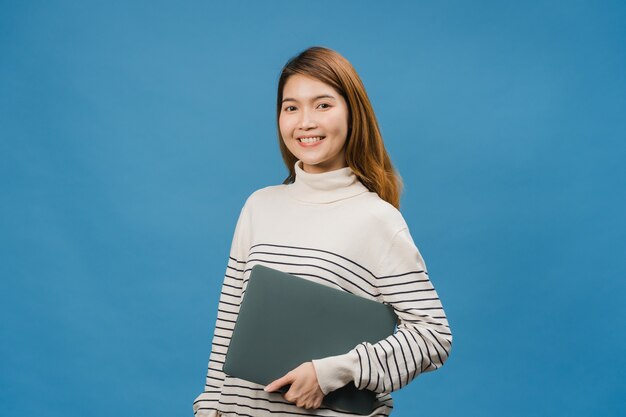 Une jeune femme asiatique surprise tient un ordinateur portable avec une expression positive, sourit largement, vêtue de vêtements décontractés et regarde devant le mur bleu