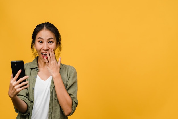 Jeune femme asiatique surprise utilisant un téléphone portable avec une expression positive, sourit largement, vêtue de vêtements décontractés et se tient isolée sur un mur jaune. Heureuse adorable femme heureuse se réjouit du succès.