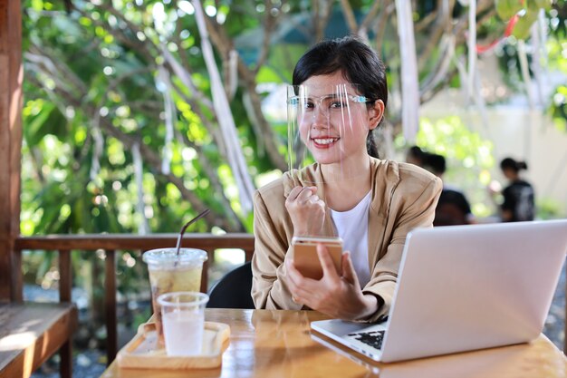 Jeune femme asiatique en tenue décontractée avec écran facial pour les soins de santé, assise dans un café et utilisant un smartphone et travaillant sur un ordinateur portable. nouveau concept normal et de distanciation sociale
