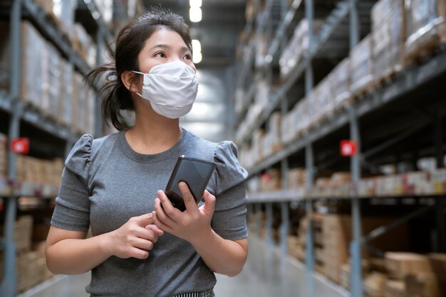 Jeune femme asiatique tenue décontractée faisant du shopping avec un chariot à chariot dans l'entrepôt d'un grand magasin avec un nouveau mode de vie normal portant une protection de masque facial et utilisant un smartphone après le verrouillage de la propagation de covid19