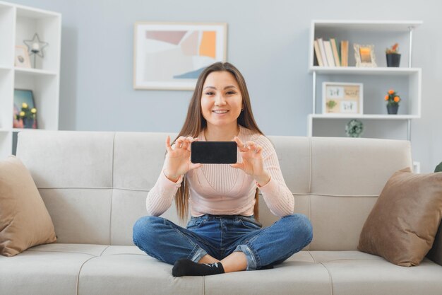 Jeune femme asiatique en vêtements décontractés assis sur un canapé à l'intérieur de la maison montrant un smartphone heureux et positif souriant amical passer du temps à la maison