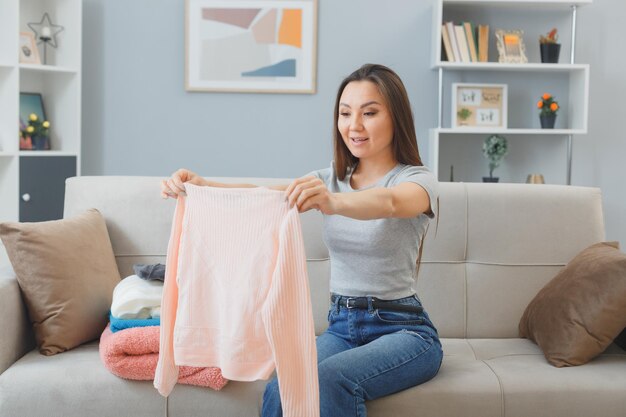 Jeune femme asiatique en vêtements décontractés assis sur un canapé avec une pile de linge propre à l'intérieur à la maison sourire heureux et positif