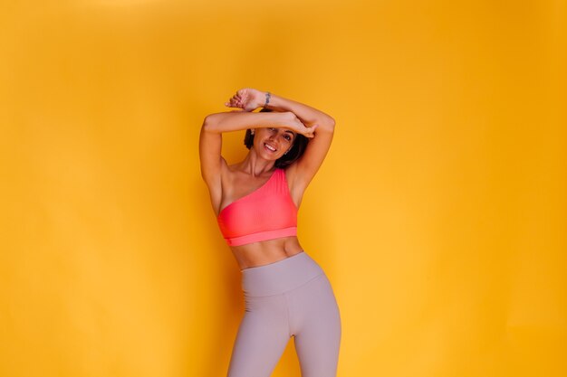 Jeune femme assez forte en forme vêtue de vêtements de sport, haut élégant et leggings, pose contre le mur jaune