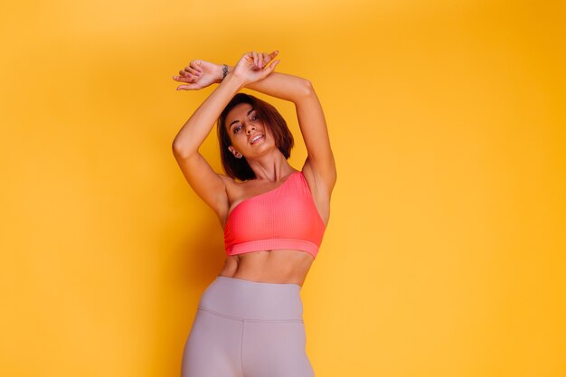 Jeune femme assez forte en forme vêtue de vêtements de sport, haut élégant et leggings, pose contre le mur jaune