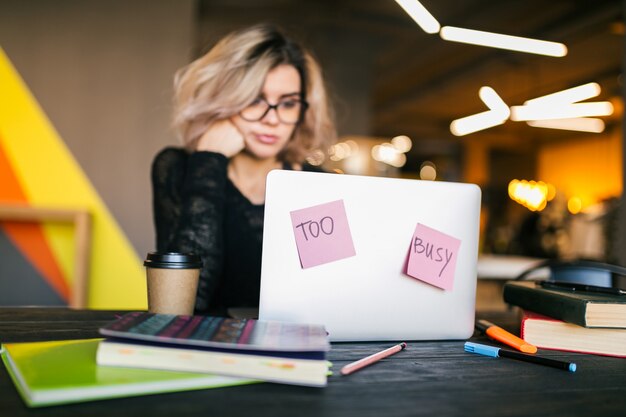 Jeune femme assez occupée assis à table travaillant sur un ordinateur portable dans un bureau de travail collaboratif, des autocollants en papier, portant des lunettes, une concentration, un étudiant en salle de classe