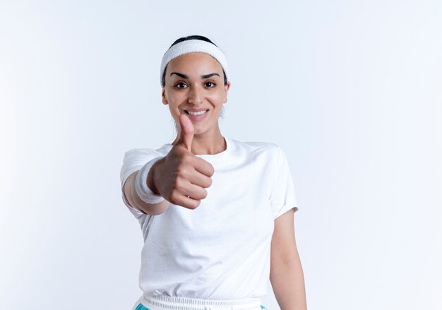 Jeune femme assez sportive souriante portant bandeau et bracelets pouces vers le haut isolé sur un espace blanc avec espace copie