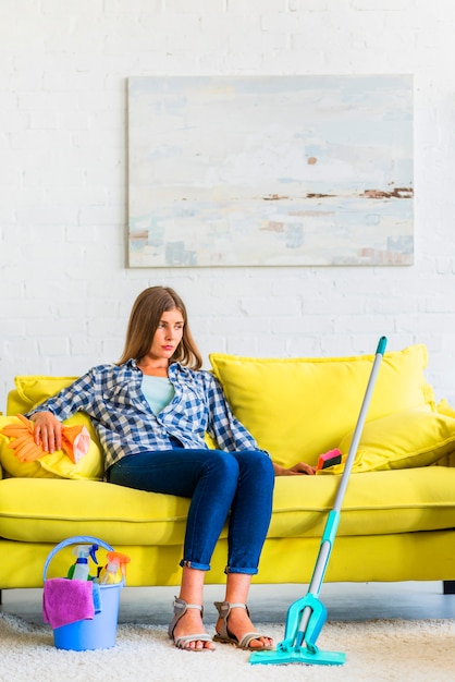 Jeune femme assise sur un canapé avec du matériel de nettoyage à la maison