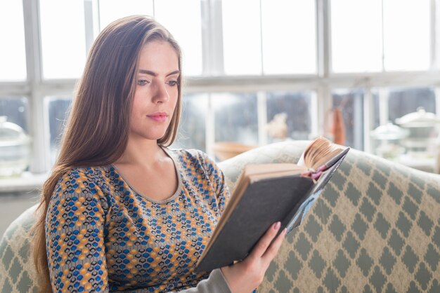 Jeune femme assise sur un canapé en lisant un livre à la maison