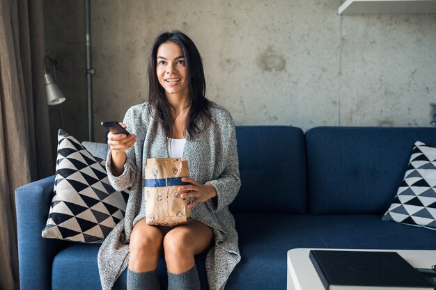 Jeune femme assise sur un canapé à la maison à regarder des films à la télévision tenant la télécommande, changer de chaîne, manger du pop-corn, s'amuser, joyeux, heureux, drôle d'expression du visage surpris, émotionnel