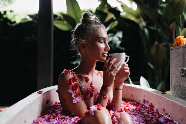 Photo gratuite jeune femme assise dans le bain les yeux fermés et boire du thé chaud. portrait de jeune fille magnifique aux cheveux blonds faisant spa et appréciant le café.