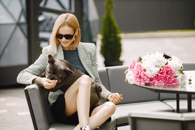 Jeune femme assise dans un café à l'extérieur et tenant un bouledogue français noir sur ses genoux. Fille portant des lunettes de soleil noires, un short et une veste grise