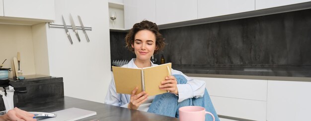 Une jeune femme assise dans la cuisine lisant ses notes dans un cahier souriant une fille à la maison faisant ses devoirs