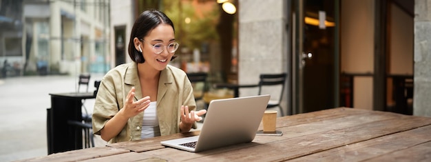 Jeune femme assise lors d'une réunion en ligne dans un café en plein air parlant à la caméra d'un ordinateur portable expliquant quelque chose