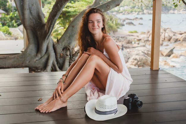 Jeune femme assise sur le sol pieds nus en robe pâle, souriant, beauté naturelle, chapeau de paille, appareil photo numérique,