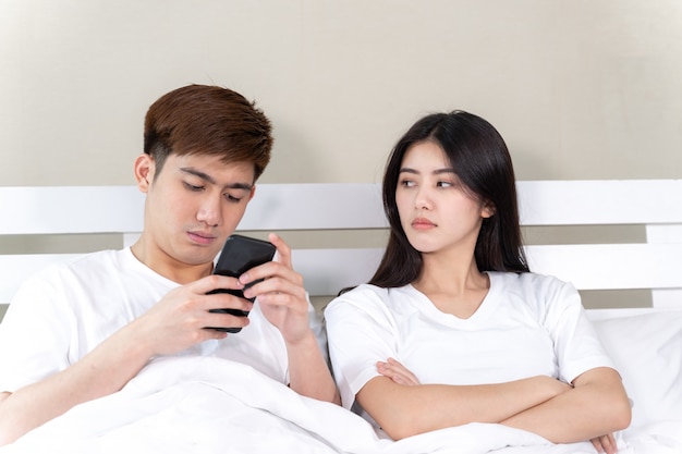 jeune femme assise sur son lit avec un sentiment de contrariété, son mari utilise un smartphone en sieste