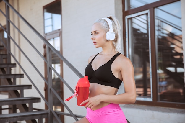 Une jeune femme athlétique dans un casque blanc travaillant à écouter de la musique dans un escalier à l'extérieur. Boire de l'eau de la bouteille de sport. Concept de mode de vie sain, sport, activité, perte de poids.