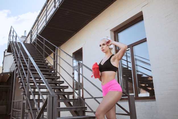 Une jeune femme athlétique dans un casque blanc travaillant à écouter de la musique dans un escalier à l'extérieur. Boire de l'eau de la bouteille de sport. Concept de mode de vie sain, sport, activité, perte de poids.