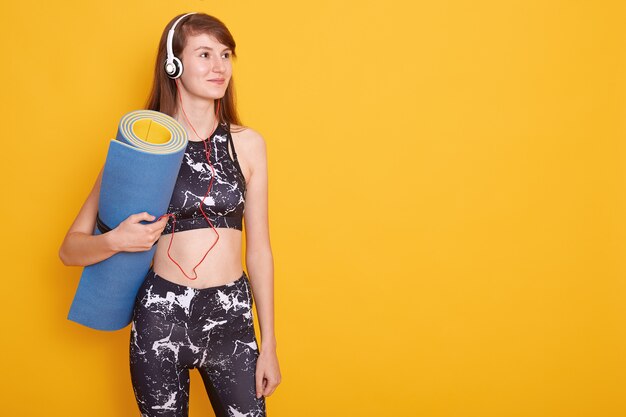 Jeune femme athlétique portant des écouteurs et des vêtements de sport noirs tenant un tapis de yoga bleu