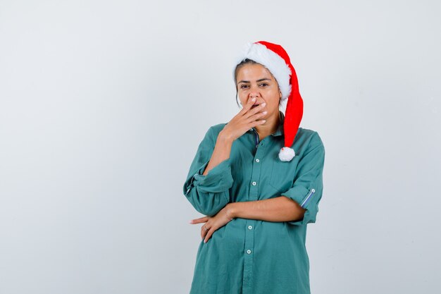 Jeune femme au chapeau de Noël, chemise avec la main sur la bouche et l'air perplexe, vue de face.