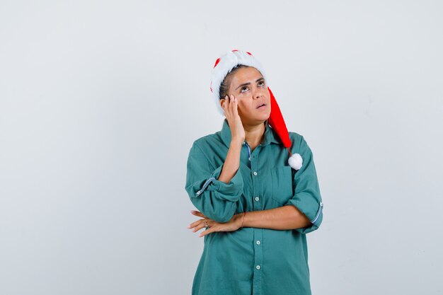 Jeune femme au chapeau de Noël, chemise avec la main près du visage et l'air pensif, vue de face.