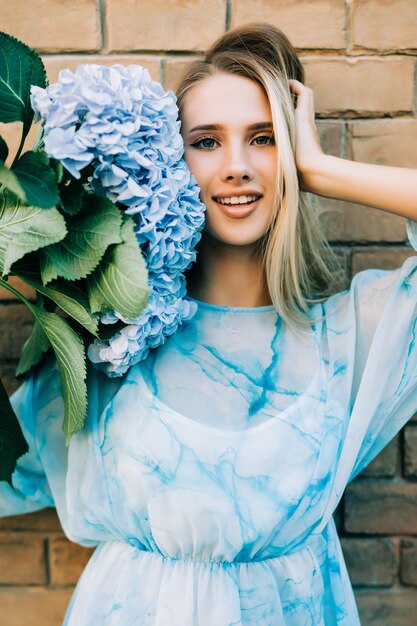 Jeune femme au chapeau de paille tenant des fleurs d'hortensia bleu debout contre le mur de la rue Breek