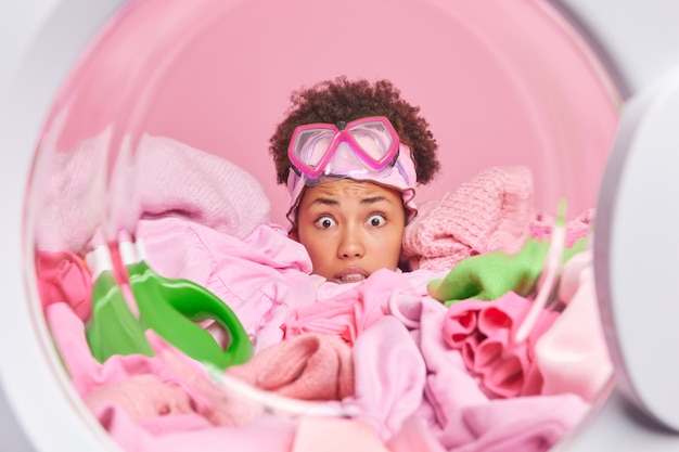 Une jeune femme au foyer inquiète abasourdie a peur que l'expression se cache dans un tas de poses de linge de l'intérieur de la laveuse porte des lunettes de plongée en apnée sur le front occupée par le travail domestique quotidien