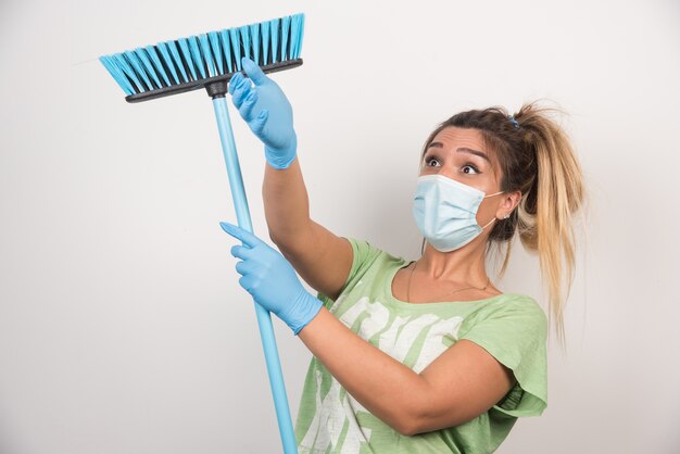 Jeune femme au foyer avec masque à faire le ménage avec un balai sur un mur blanc.