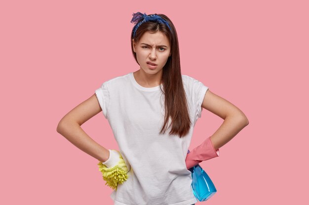 Une jeune femme au foyer surmenée se sent fatiguée, garde les mains sur la taille, porte un t-shirt décontracté, un bandeau, des gants en caoutchouc, a l'air mécontent