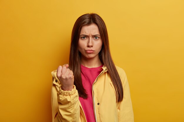 Une jeune femme autoritaire montre le poing fermé, regarde en colère, promet de se venger d'un mauvais comportement, porte un coupe-vent, isolé sur un mur jaune. Je vais te montrer. Dame menace à l'intérieur