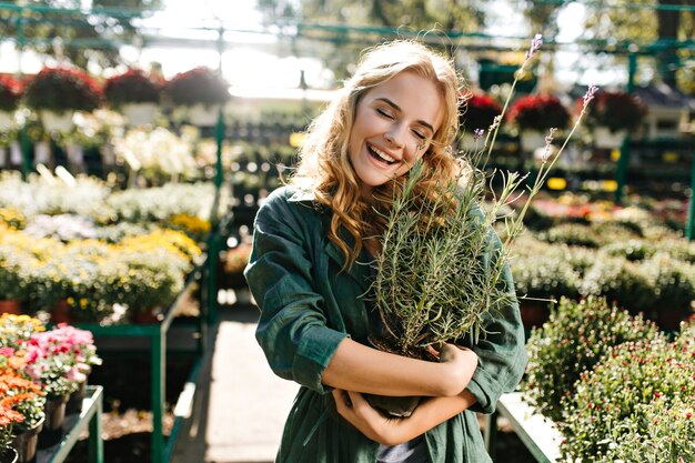 Jeune femme aux beaux cheveux blonds et sourire doux, vêtue d'une robe verte avec ceinture travaille en serre