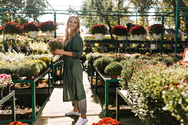 Jeune femme aux beaux cheveux blonds et sourire doux, vêtue d'une robe verte avec ceinture travaille en serre