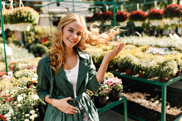 Jeune femme aux beaux cheveux blonds et sourire doux, vêtue d'une robe verte avec ceinture travaille en serre