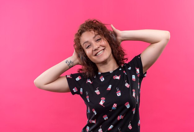 Jeune femme aux cheveux bouclés courts avec un casque sur la tête appréciant sa musique préférée happy smiling debout sur le mur rose