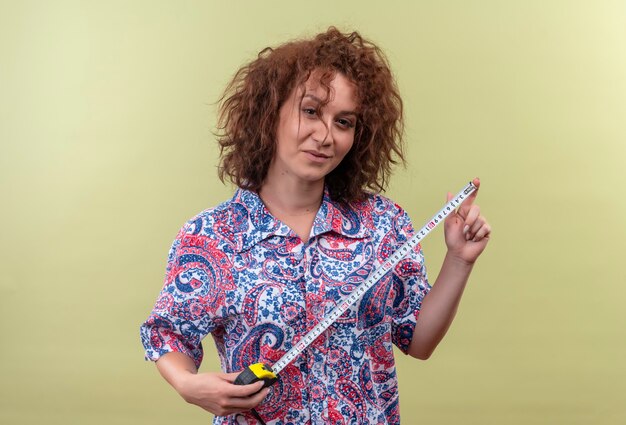 Jeune femme aux cheveux bouclés courts en chemise colorée à l'aide de ruban à mesurer smiling confiant debout sur mur vert