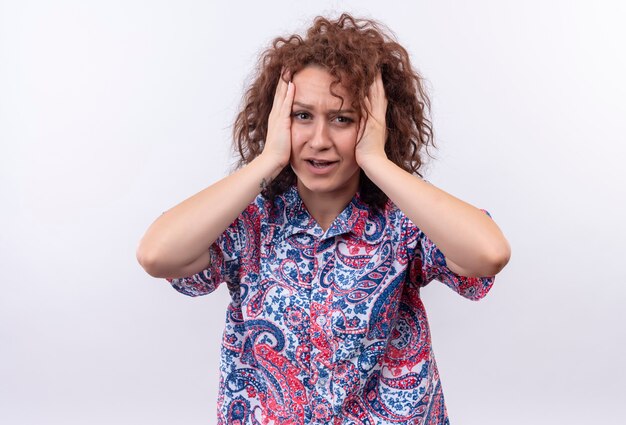 Jeune femme aux cheveux bouclés courts en chemise colorée choquée de toucher sa tête debout sur un mur blanc